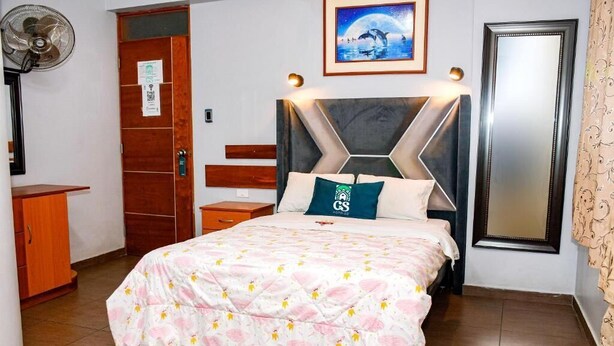 Une chambre d’hôtel comprenant un lit, une armoire en bois, une table de chevet, un ventilateur fixé au mur, un tableau encadré et un miroir.