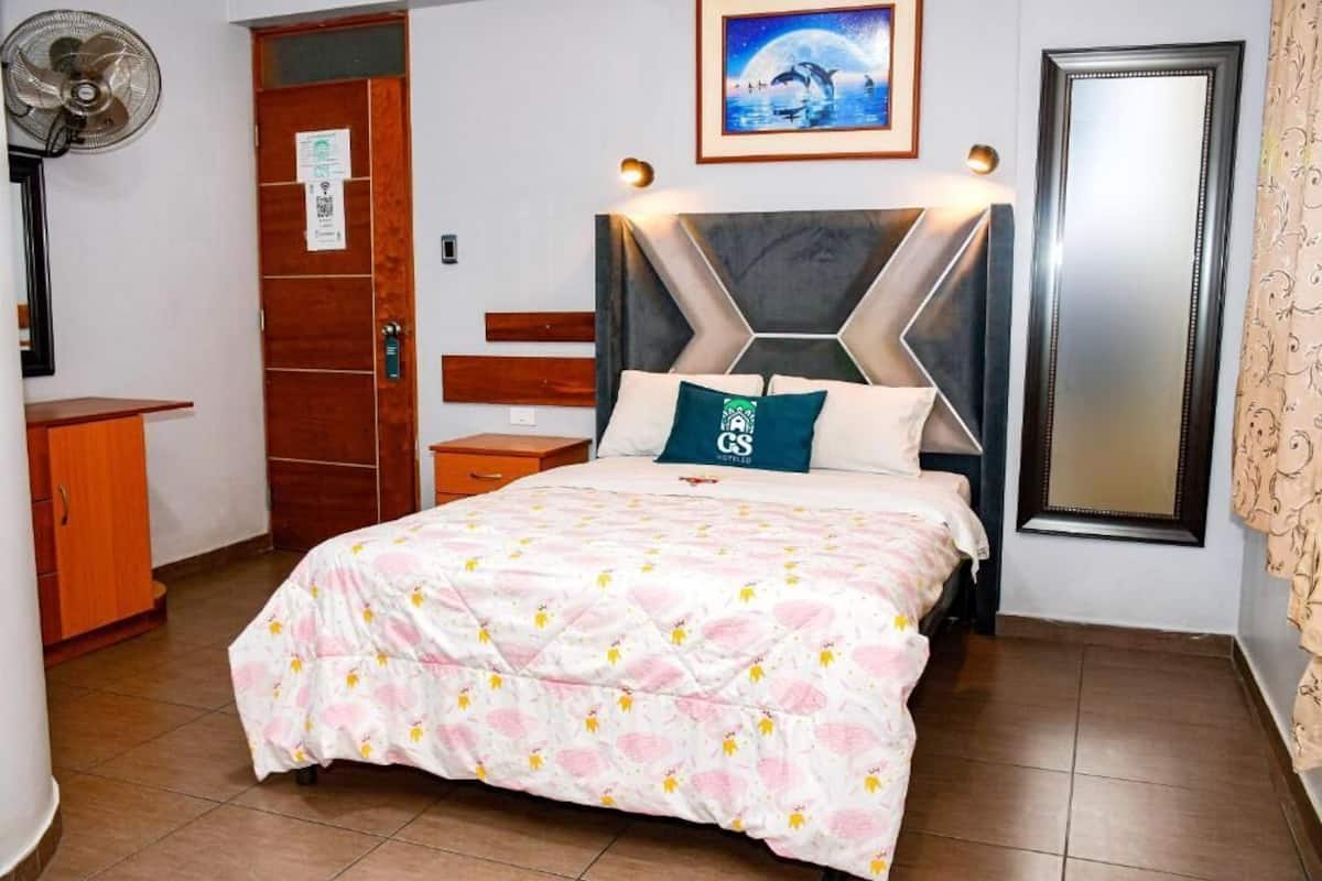 A hotel room with a bed, a wooden wardrobe, a nightstand, a wall-mounted fan, a framed picture, and a mirror.
