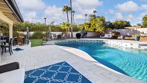 A swimming pool with a tiled edge, surrounded by a patio area with outdoor furniture and palm trees.