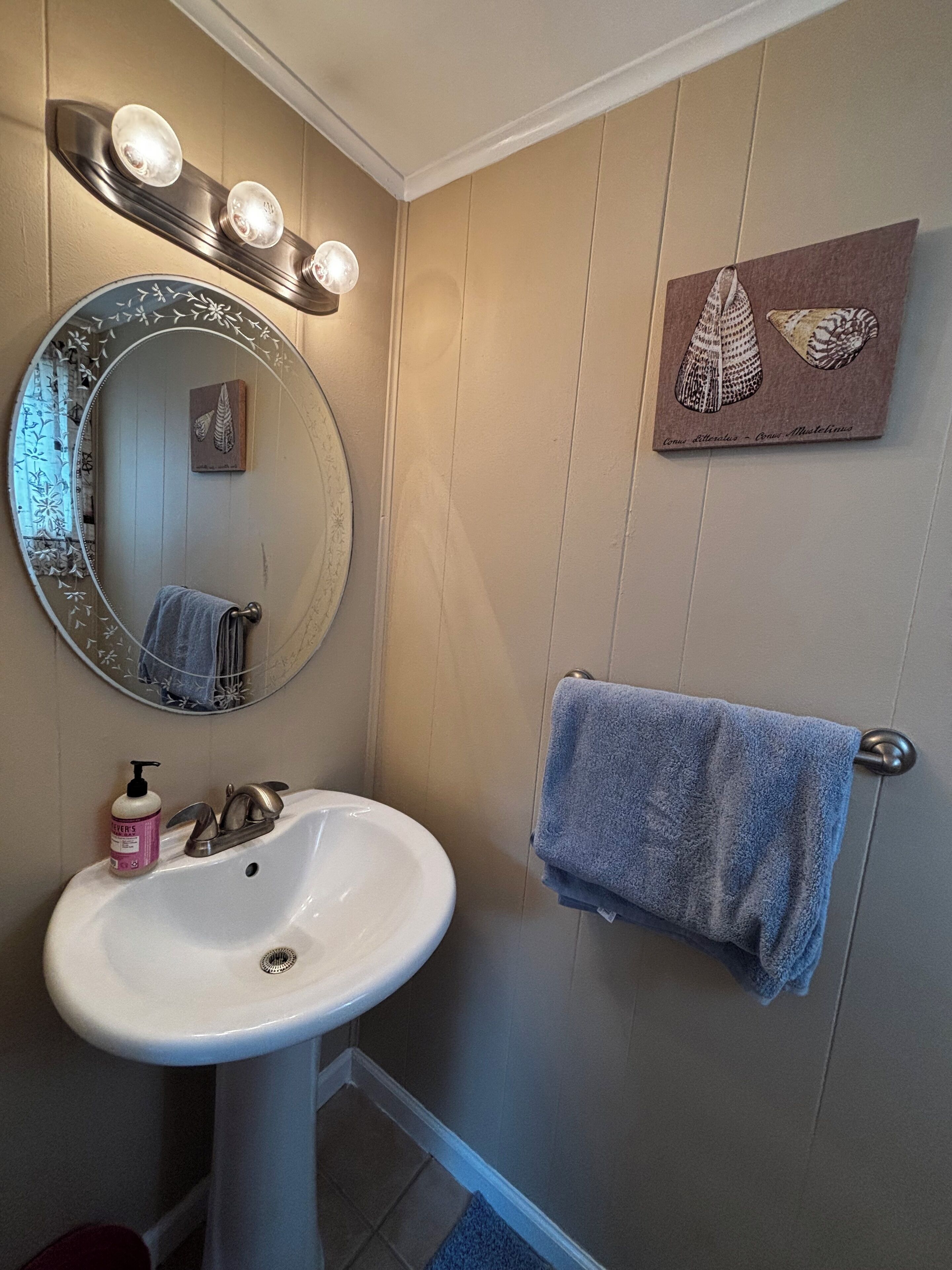 Une salle de bain avec un lavabo blanc, un miroir rond avec éclairage, un porte-serviettes et un tableau encadré au mur.