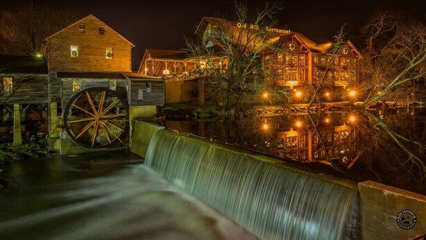 Eine historische Wassermühle mit einem großen hölzernen Wasserrad, ein Holzhaus mit einem Schild, auf dem 