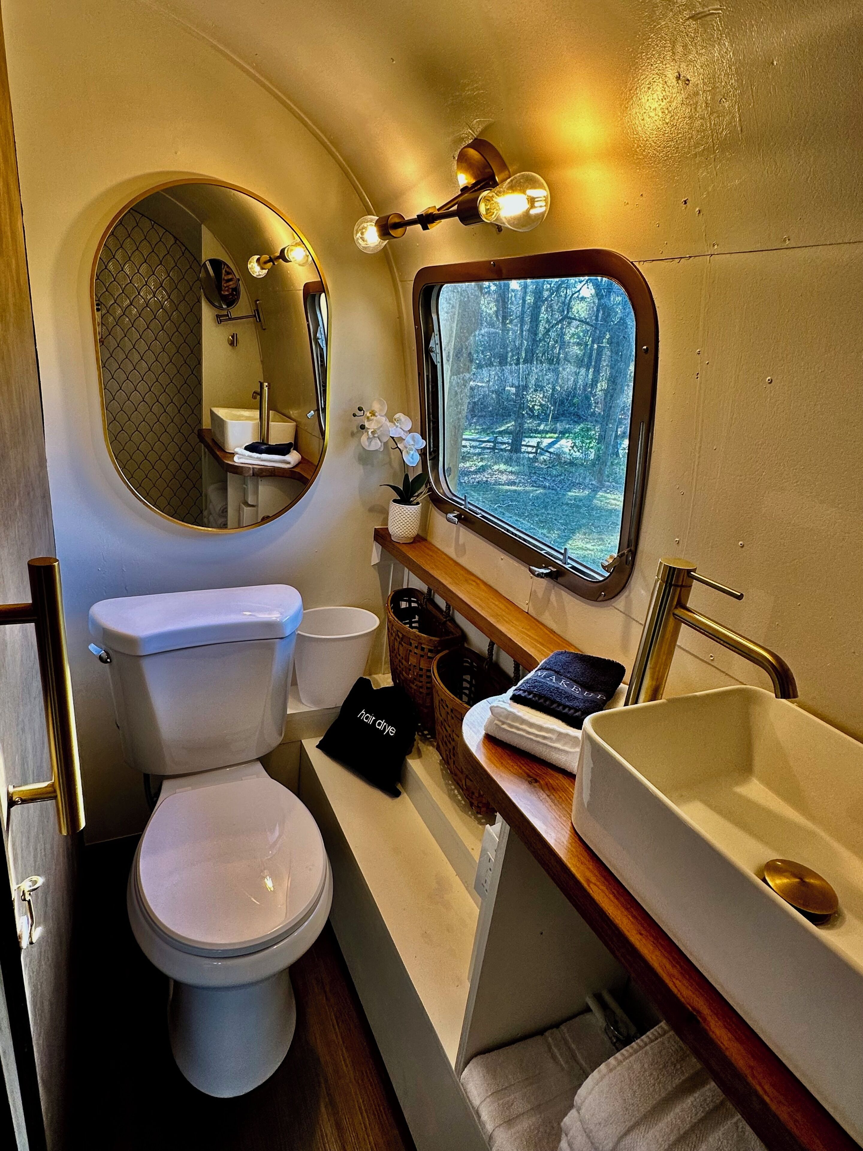 A compact bathroom with a toilet, sink, and mirror. A window provides a view of a forested area.