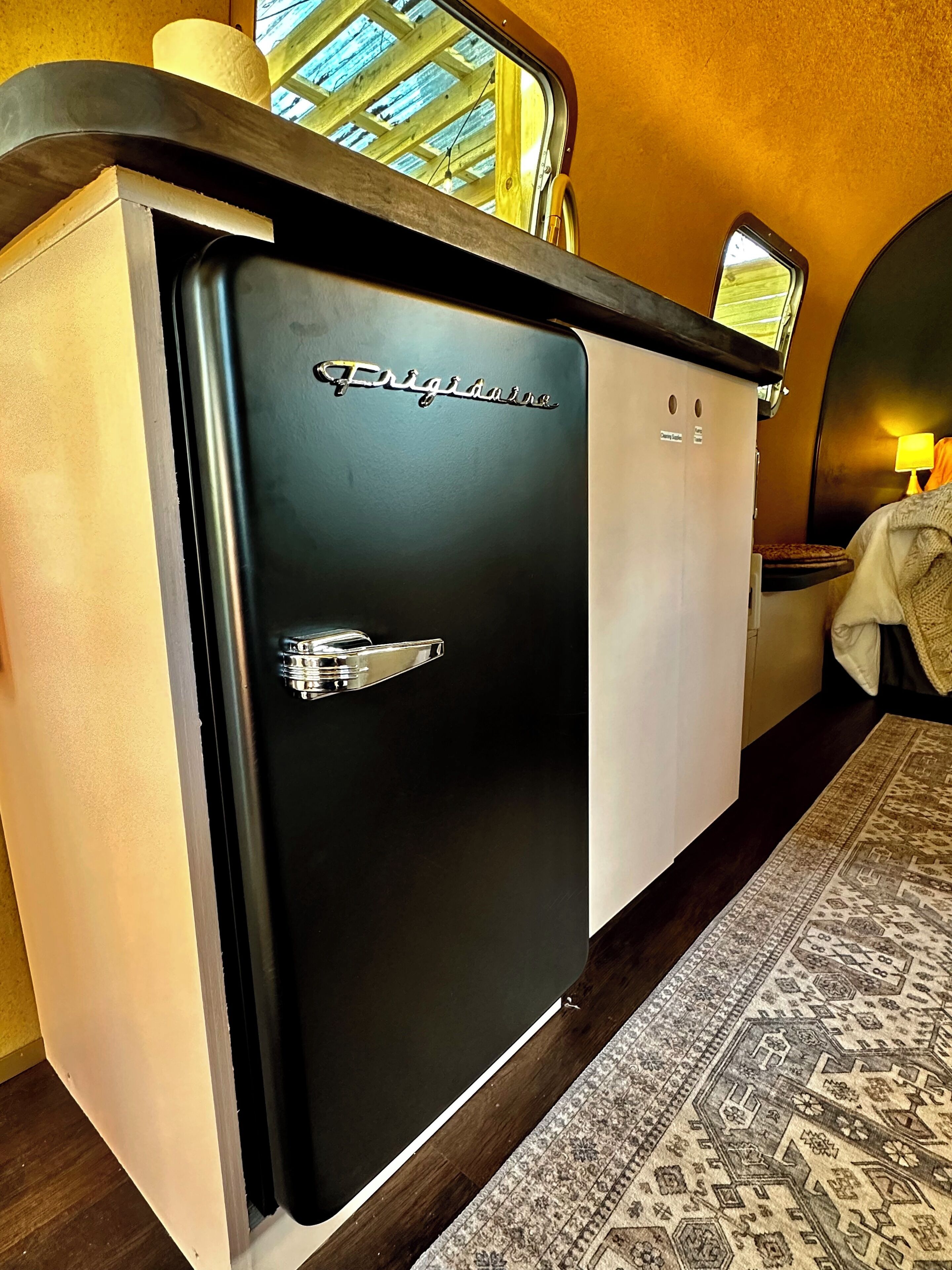 A compact, modern kitchenette with a black refrigerator labeled "Frigidaire," a small countertop, and a window above the sink.