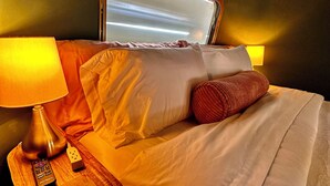 Una cama bien hecha con sábanas blancas, una mesita de noche de madera con una lámpara, y un espejo sobre la cama.