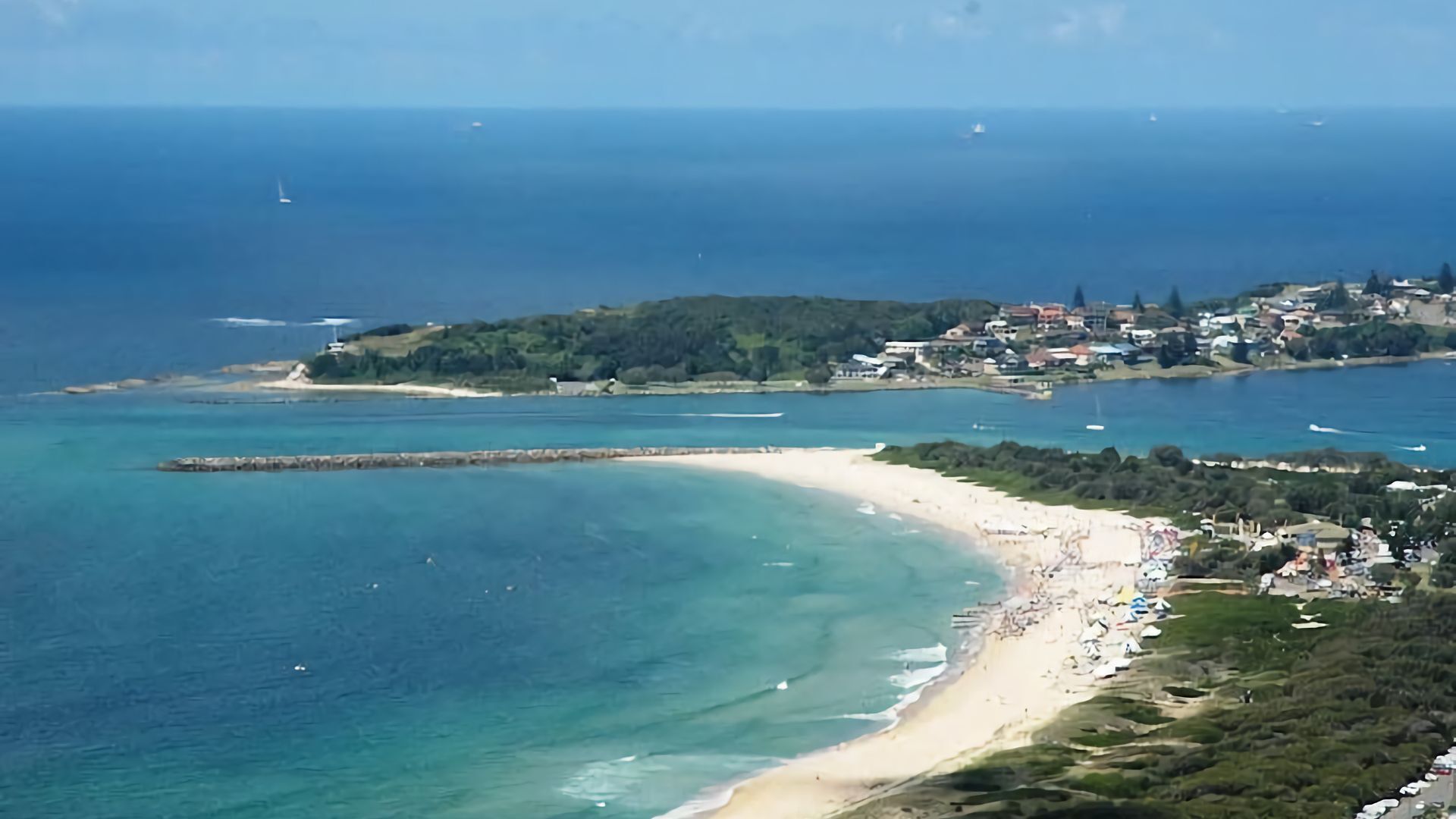 A coastal town with a sandy beach, clear blue waters, and a hilly backdrop.