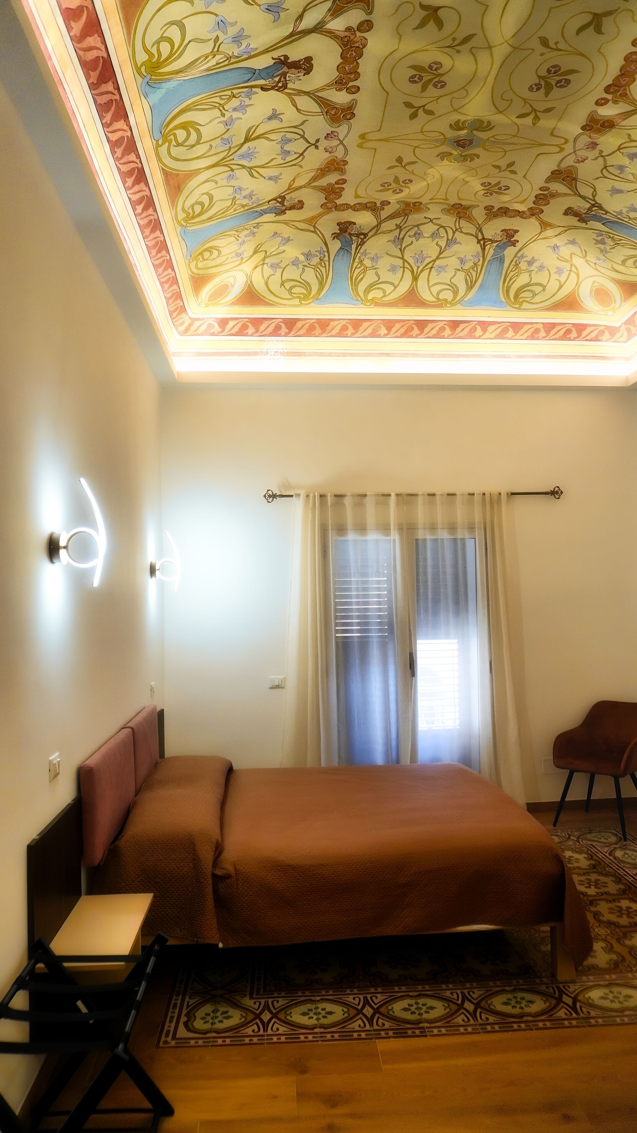 Un dormitorio con una cama, una silla, un ventanal con cortinas y un techo decorativo.