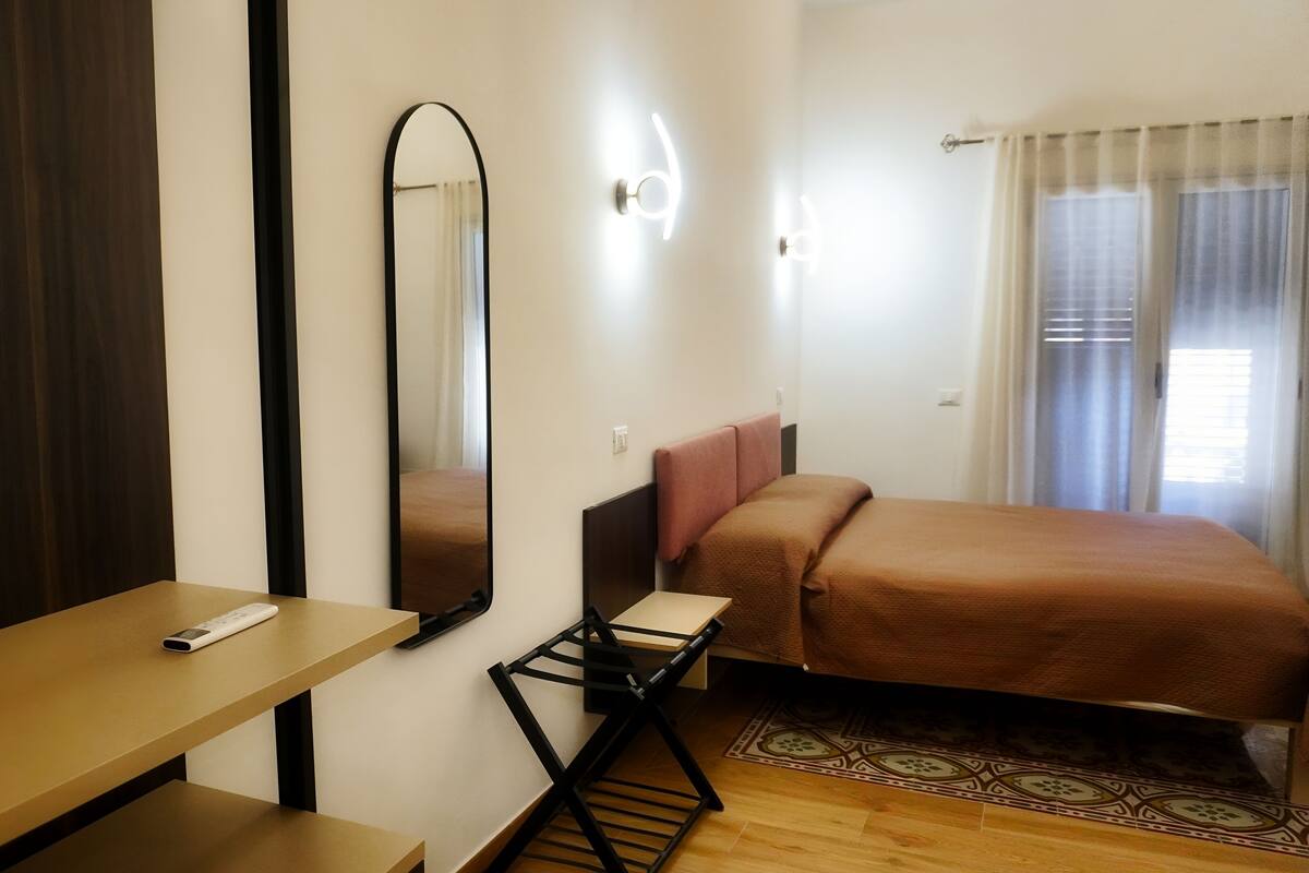 A hotel room with a bed, a desk, a chair, a mirror, and a window with curtains.