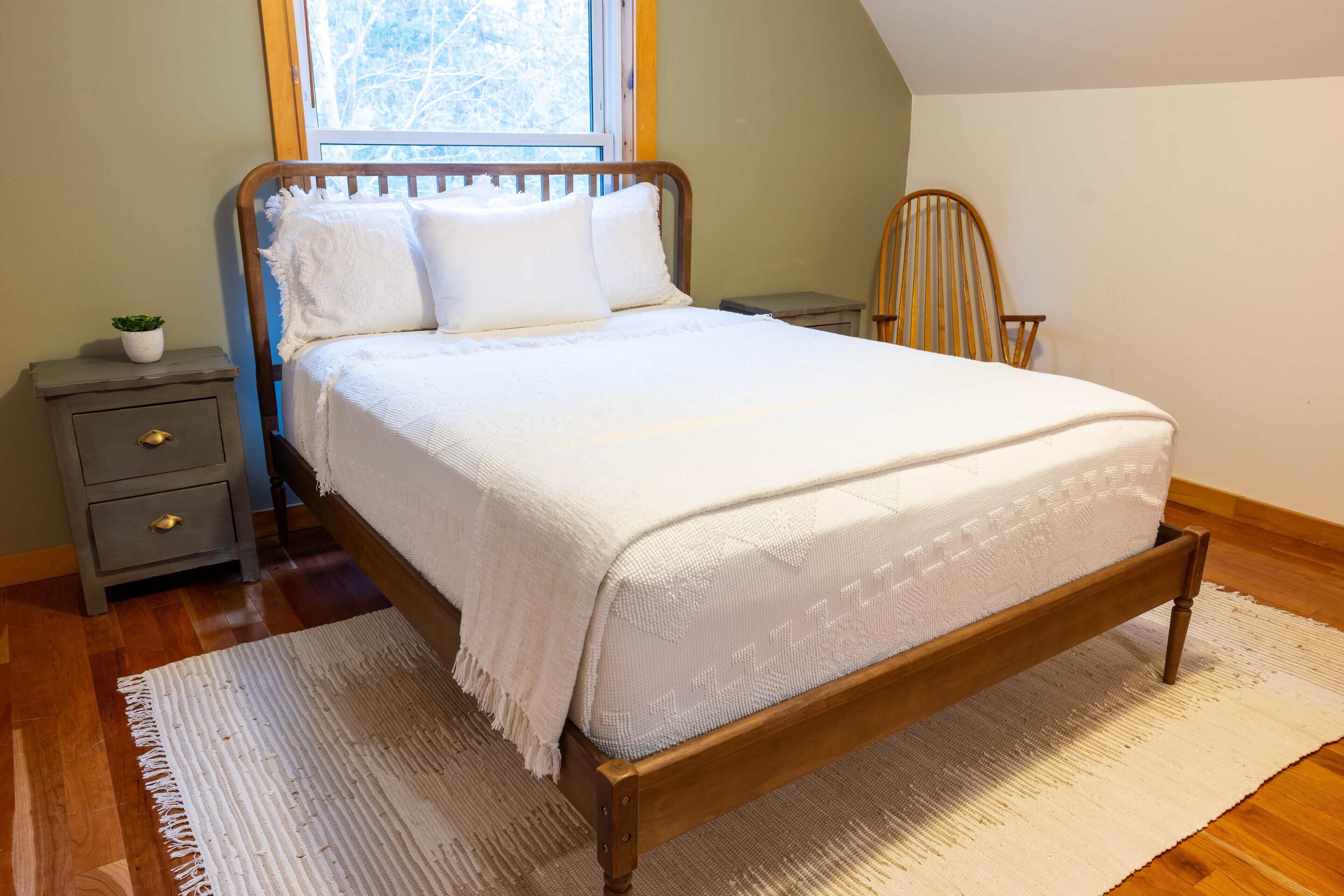 A neatly made bed with a wooden frame and white bedding, a small bedside table with a plant, a chair, and a window with a view of trees.