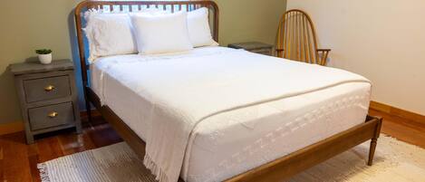 A neatly made bed with a wooden frame and white bedding, a small bedside table with a plant, a chair, and a window with a view of trees.