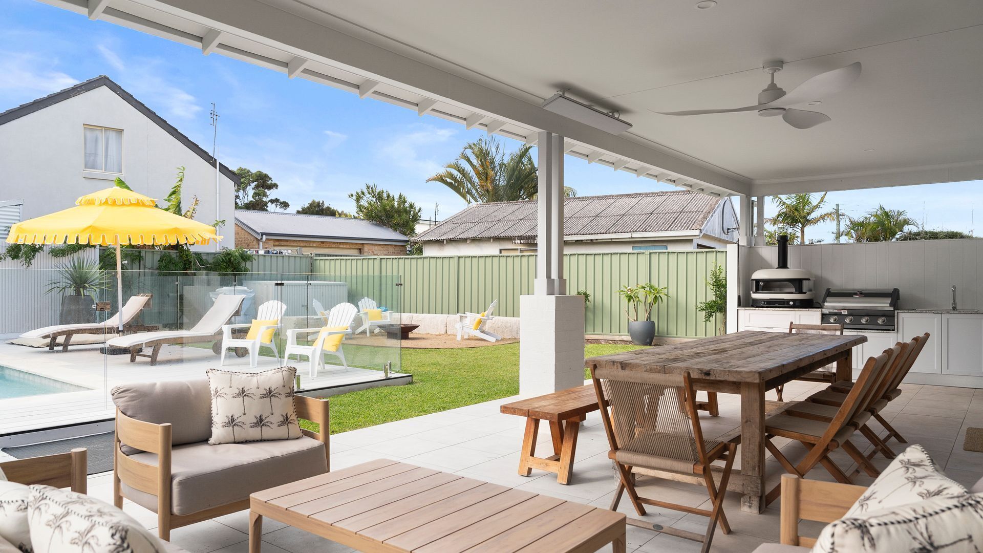 A covered outdoor patio with a pool, lounge chairs, a wooden table, and a barbecue grill.