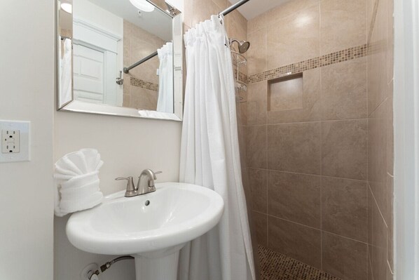 Un baño con lavamanos blanco, espejo y ducha con azulejos color beige.
