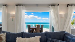 Una habitación con vistas al océano, un sofá con cojines, una mesa con sillas y cortinas blancas.