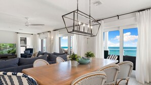 Amplia sala de estar con una gran mesa de comedor de madera, sillas de mimbre y un sofá. Se ve el océano a través de ventanas grandes.