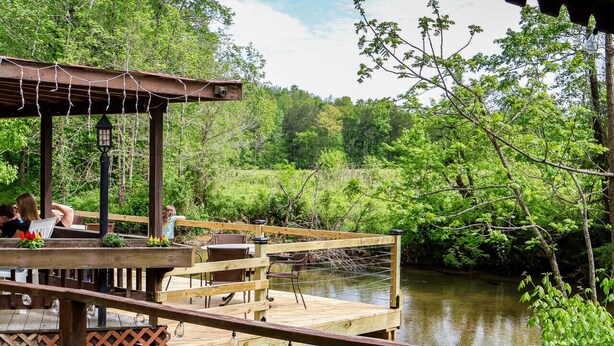 Een houten terras met stoelen en tafels, uitkijkend op een rivier en omgeven door groen.