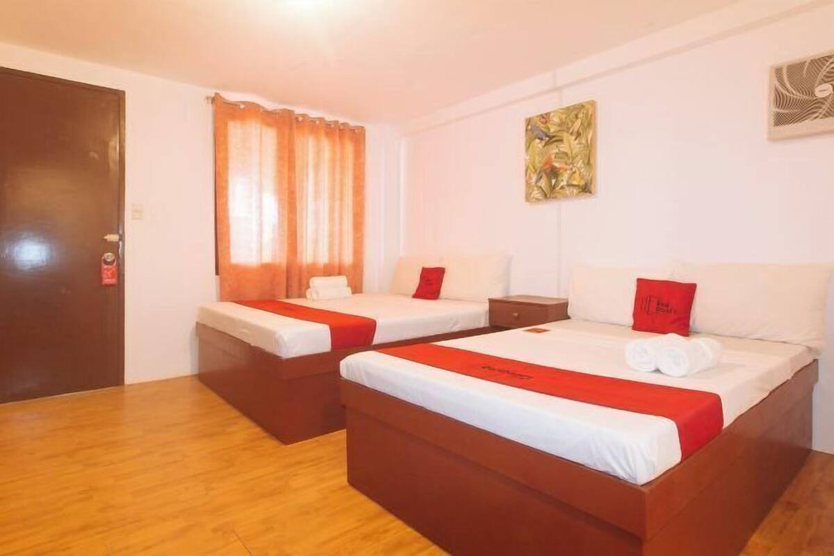 A hotel room with two beds, a wooden floor, a door, and a window with curtains.
