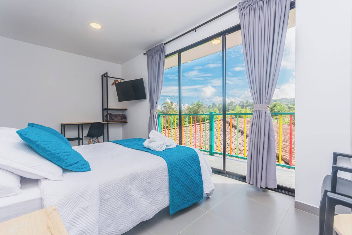 A hotel room with a bed, a TV, and a balcony with a view of greenery and a fence.