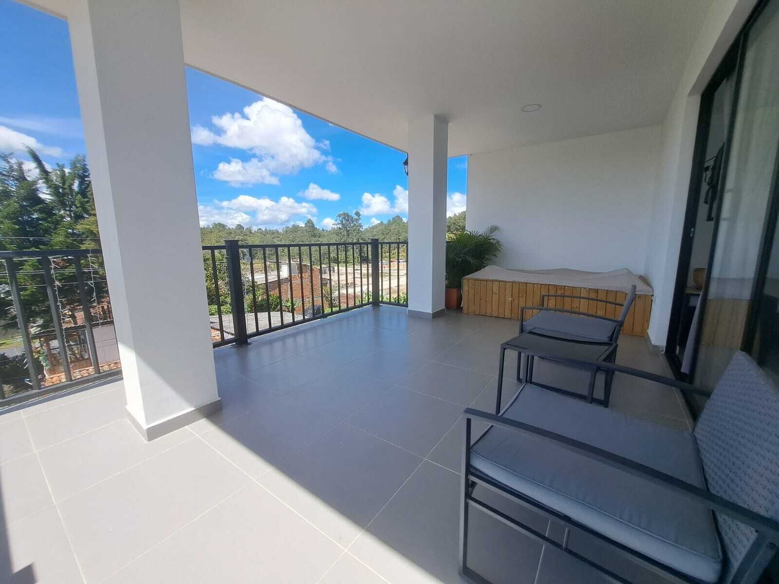 A balcony with a view of a green landscape and a clear sky. There is a wooden bench and a table with chairs.
