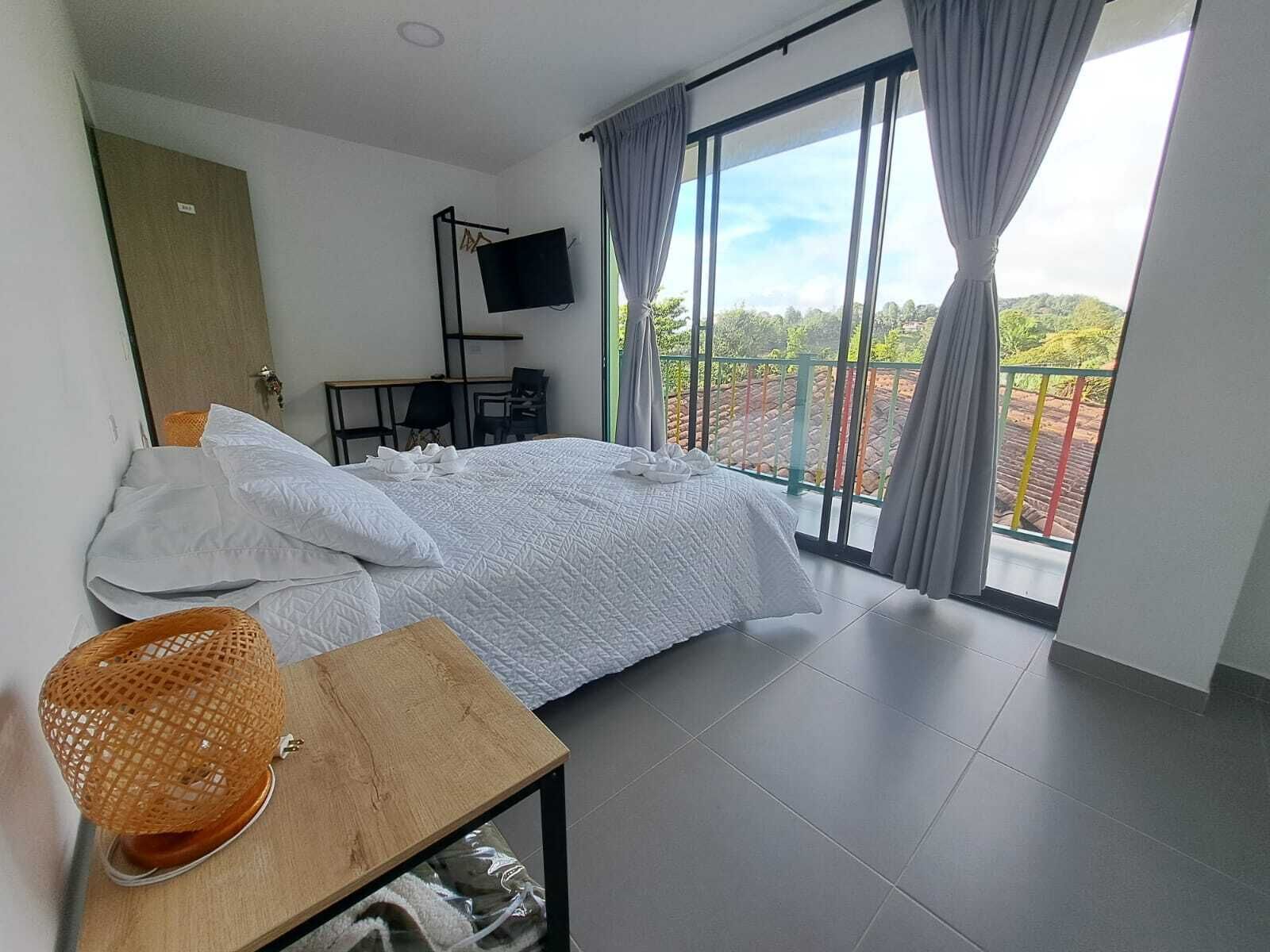 Quarto de hotel com cama, televisão, escrivaninha e vista para uma varanda com guarda-corpo.