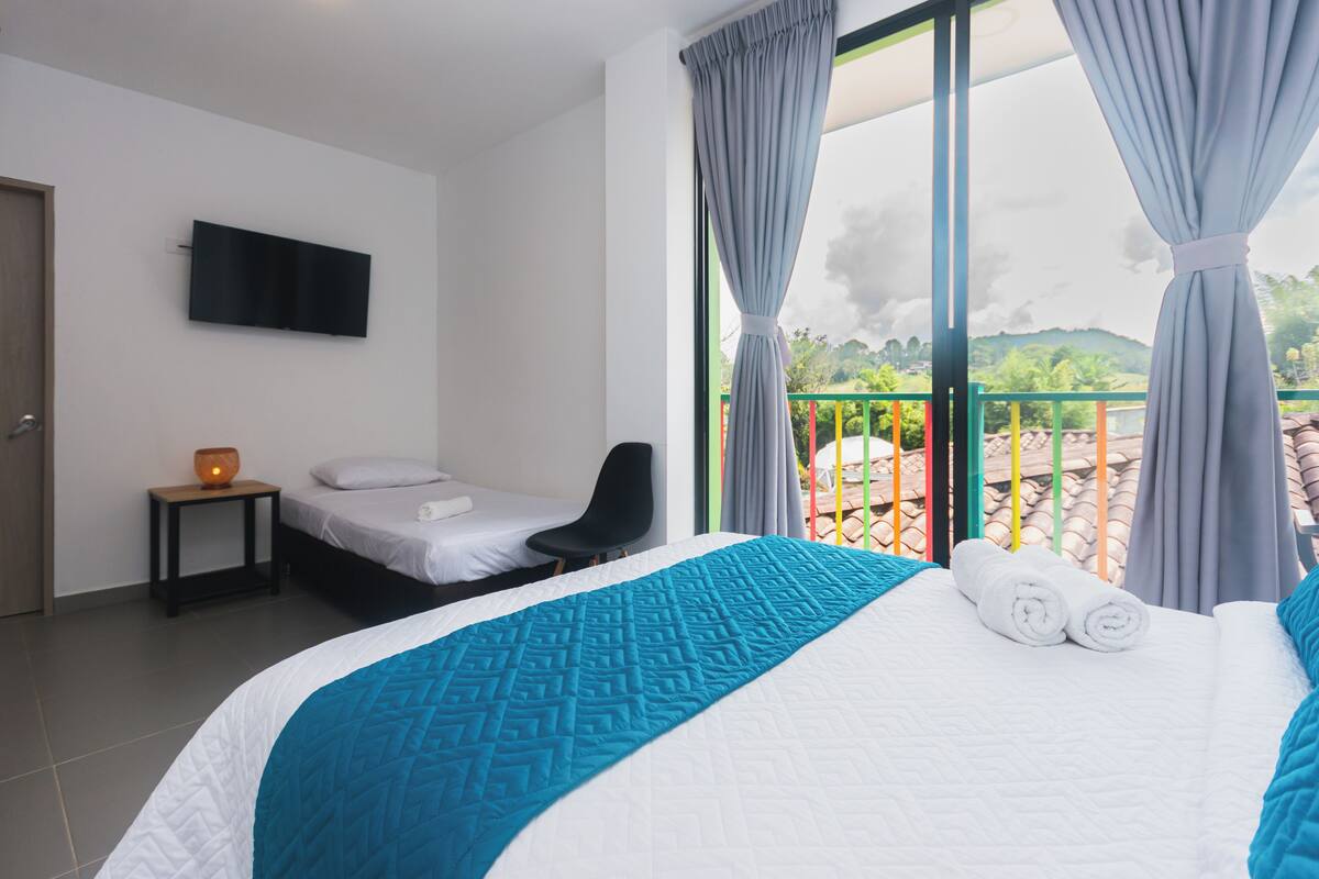 A hotel room with a large bed, a TV mounted on the wall, a small table with a lamp, and a balcony with a view of greenery and buildings.