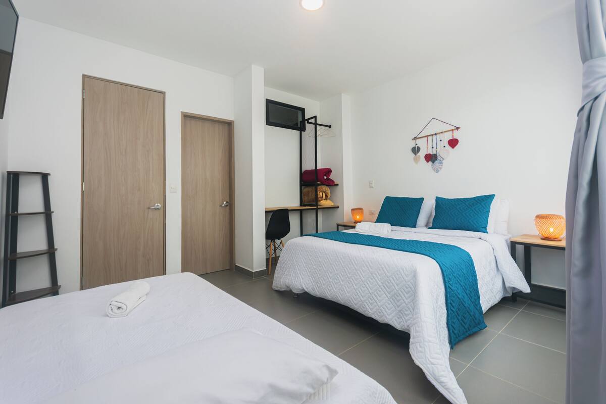 A hotel room with two beds, a desk, a chair, a wardrobe, and a wall-mounted shelf with decorative items.