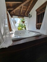 Un área de spa con una bañera de hidromasaje independiente, vigas de techo de madera y vistas a la vegetación exterior.