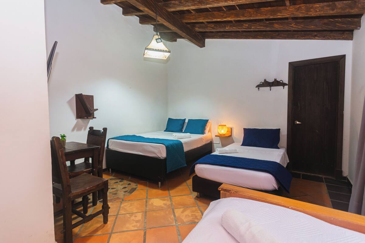 A small, cozy room with two beds, a wooden table, and a chair. There is a door and a wall-mounted shelf.