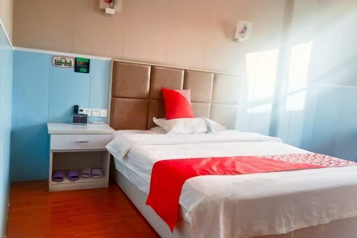 A hotel room with a bed, a nightstand, a shelf with slippers, and a wall with a sign.