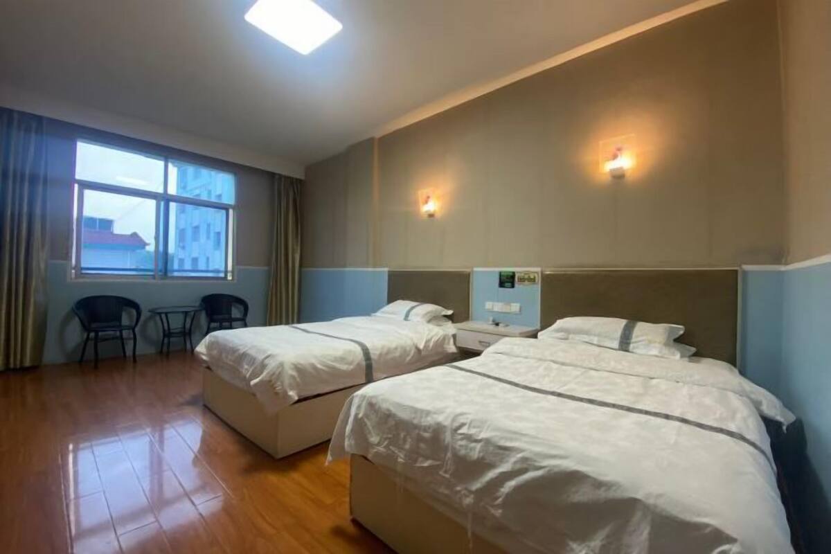 A hotel room with two beds, a small table, two chairs, a window, and a wall-mounted light.