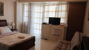 A hotel room with a bed, a TV on a stand, a chair, and a window with curtains. - Palm Tree Genlex Condo (Pasay)