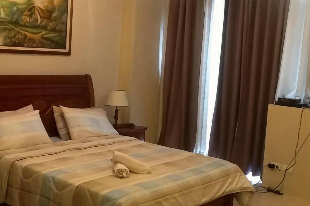 Een hotelkamer met een bed, een nachtkastje met een lamp, een schilderij aan de muur en een raam met gordijnen.