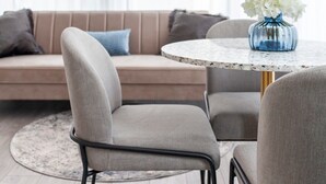 En moderne stue med et rundt bord, to grå stole, en beige sofa med puder og en vase med hvide blomster.