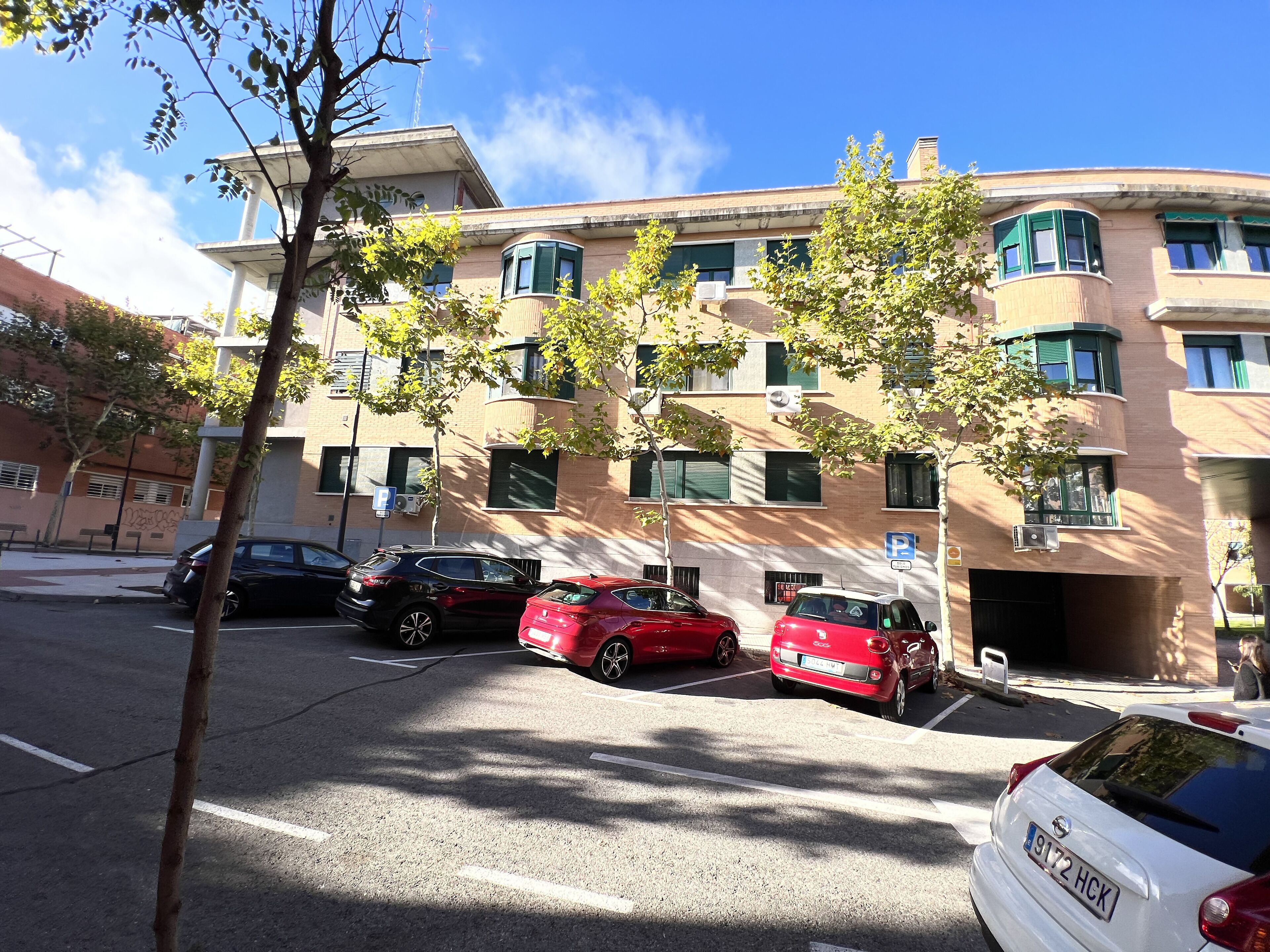 Un edificio a più piani con un parcheggio. Si notano diverse auto parcheggiate, tra cui veicoli rossi e bianchi.