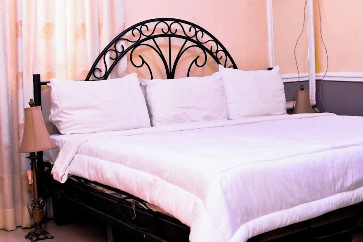 A neatly made bed with white linens and a black metal headboard in a room with a curtain and a lamp.