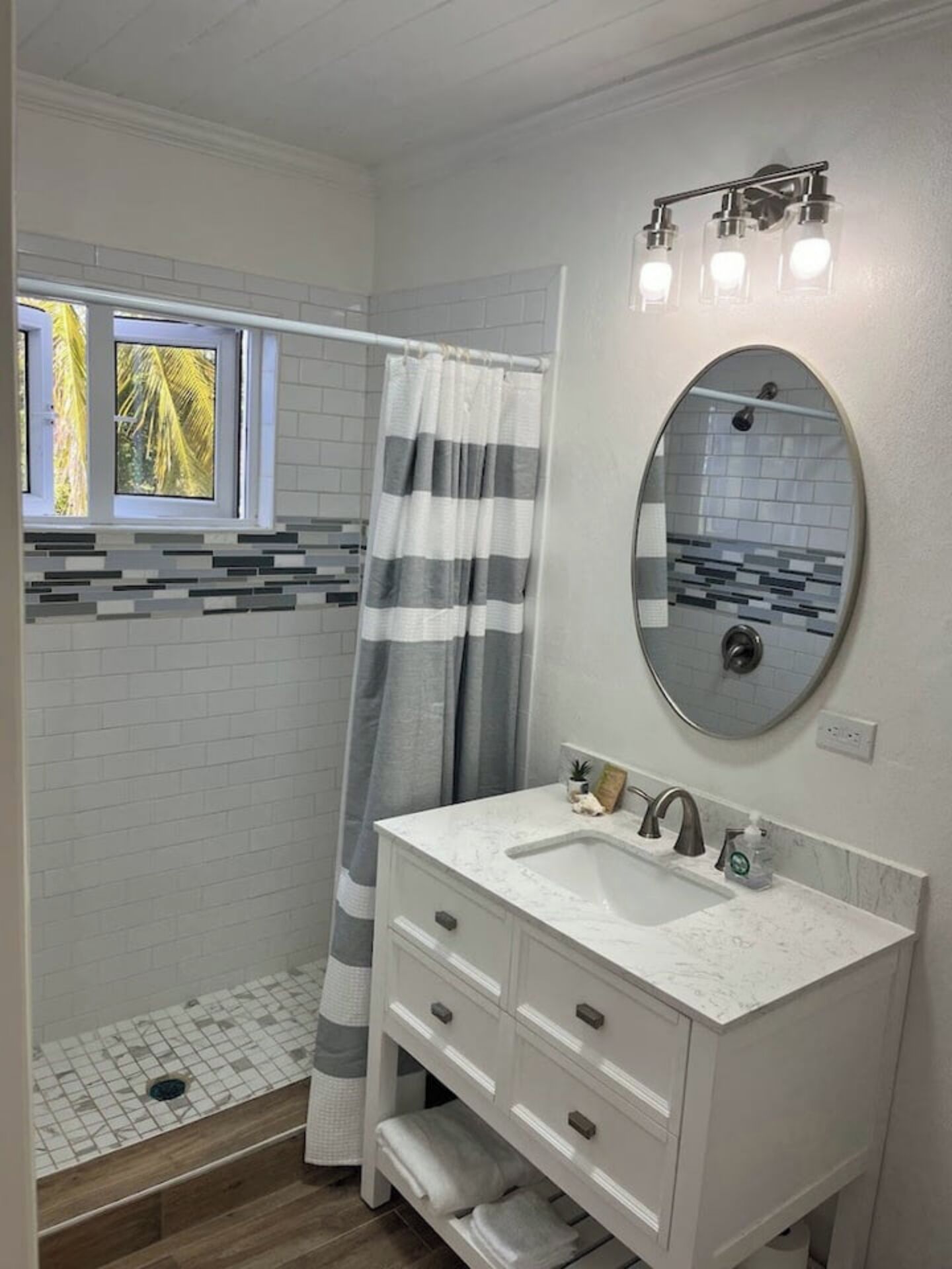A bathroom with a shower, a vanity with a sink, a mirror, and a window with a view of palm trees.