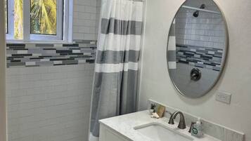A bathroom with a shower, a vanity with a sink, a mirror, and a window with a view of palm trees.