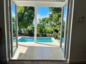 A view through a glass door of a swimming pool surrounded by lush greenery.