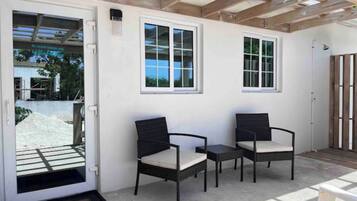 Un área de estar al aire libre con dos sillas de mimbre y una mesa pequeña, una pérgola de madera encima y una puerta blanca con felpudo.