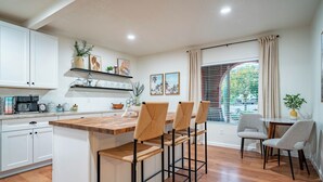 A modern kitchen with white cabinets, a wooden island, and bar stools. There are shelves with decorative items and a window with curtains. - Desert Skies - Beautiful private Tucson home with great mountain views &  pool (Tucson)