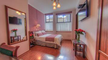 Un dormitorio ordenado con cama, mesitas de noche, un espejo y un televisor montado en la pared.