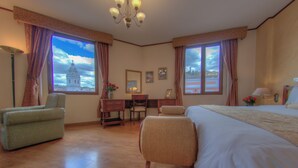 A hotel room with a bed, two armchairs, a desk, and a chair. There are two windows with curtains, a chandelier, and a view of a building. - Real Audiencia Hotel  (Quito)