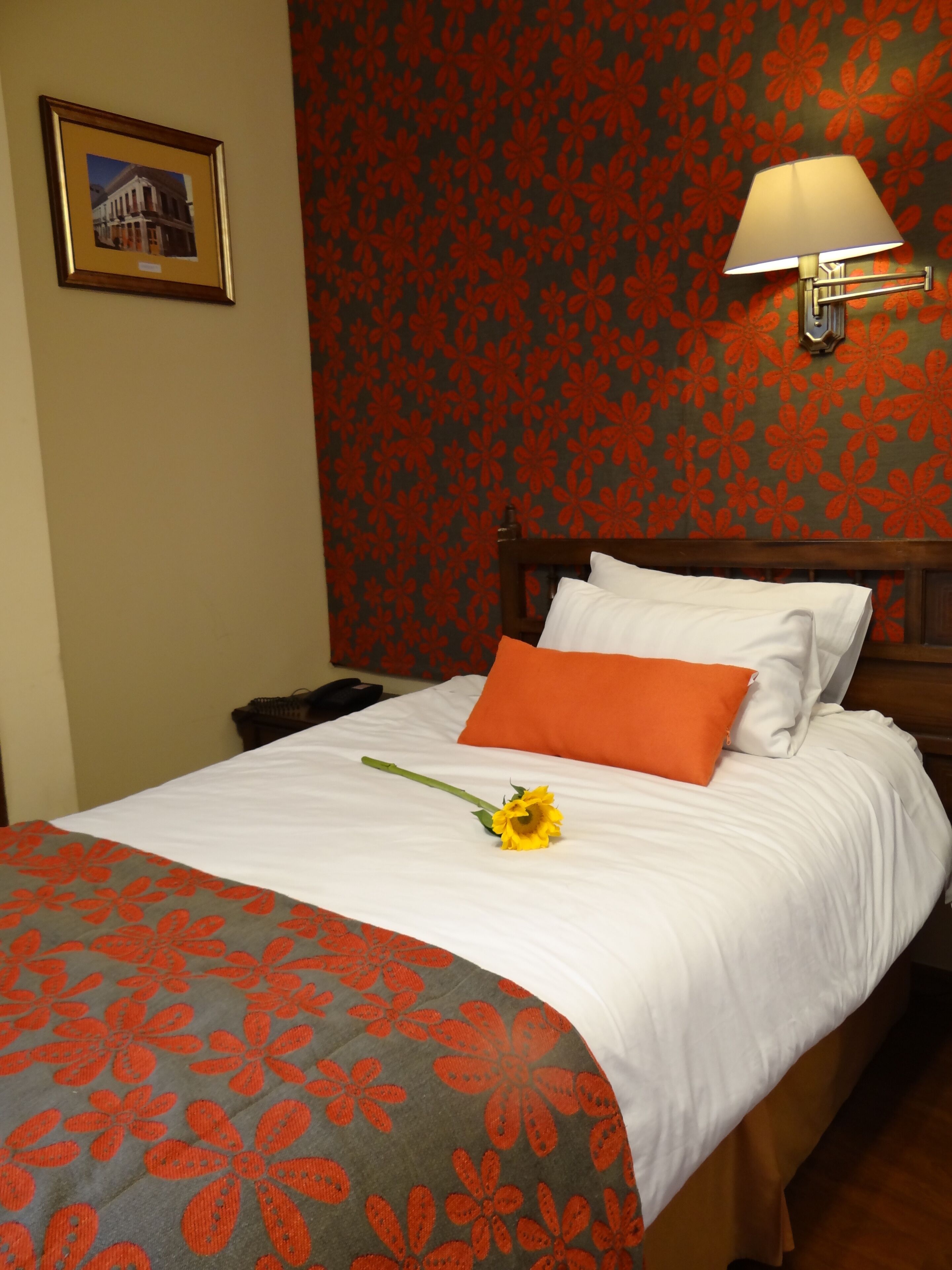 A neatly made bed with white linens, an orange pillow, and a single yellow flower.