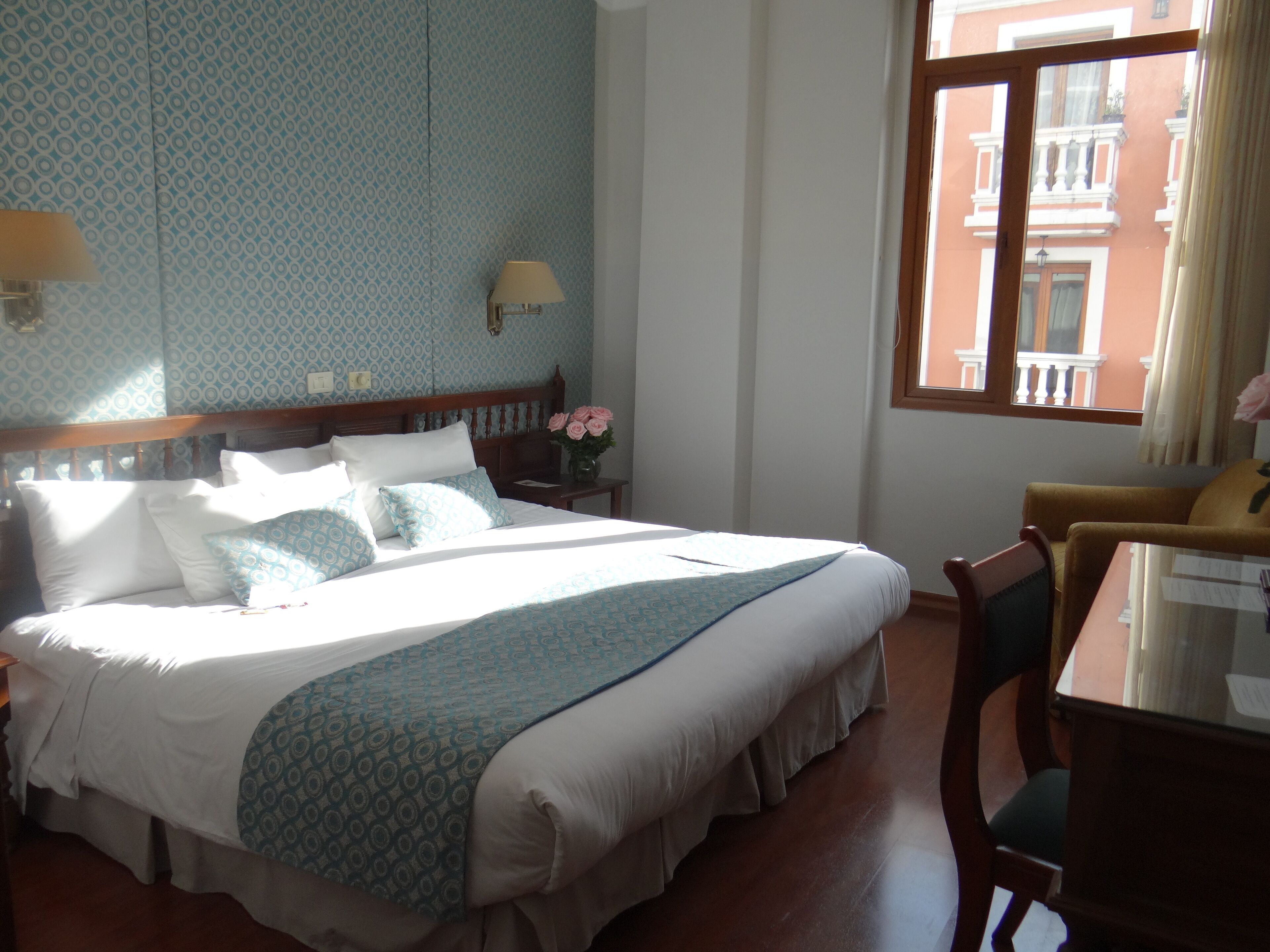 A hotel room with a large bed, a bedside table with a lamp, a vase with flowers, and a window with a view of buildings.