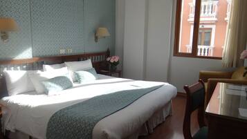 Et hotelværelse med en stor seng, et natbord med en lampe, en vase med blomster og et vindue med udsigt til bygninger.