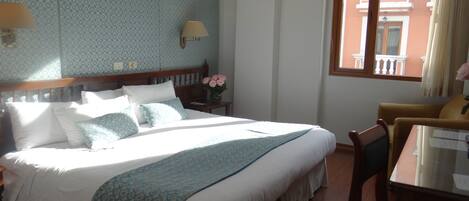 A hotel room with a large bed, a bedside table with a lamp, a vase with flowers, and a window with a view of buildings.
