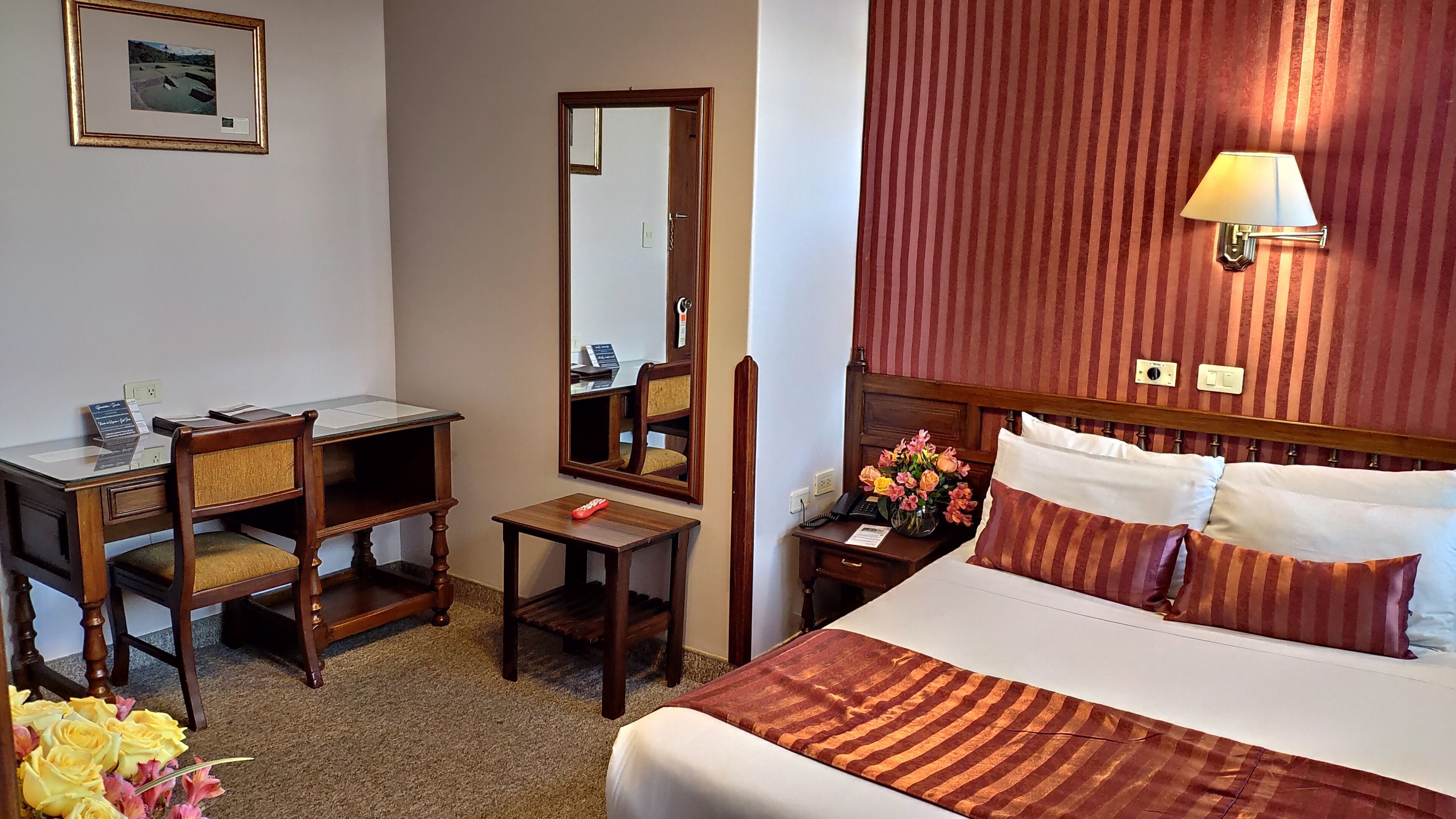 A hotel room with a bed, a desk, a chair, a mirror, a nightstand with a lamp, and a wall-mounted light fixture.