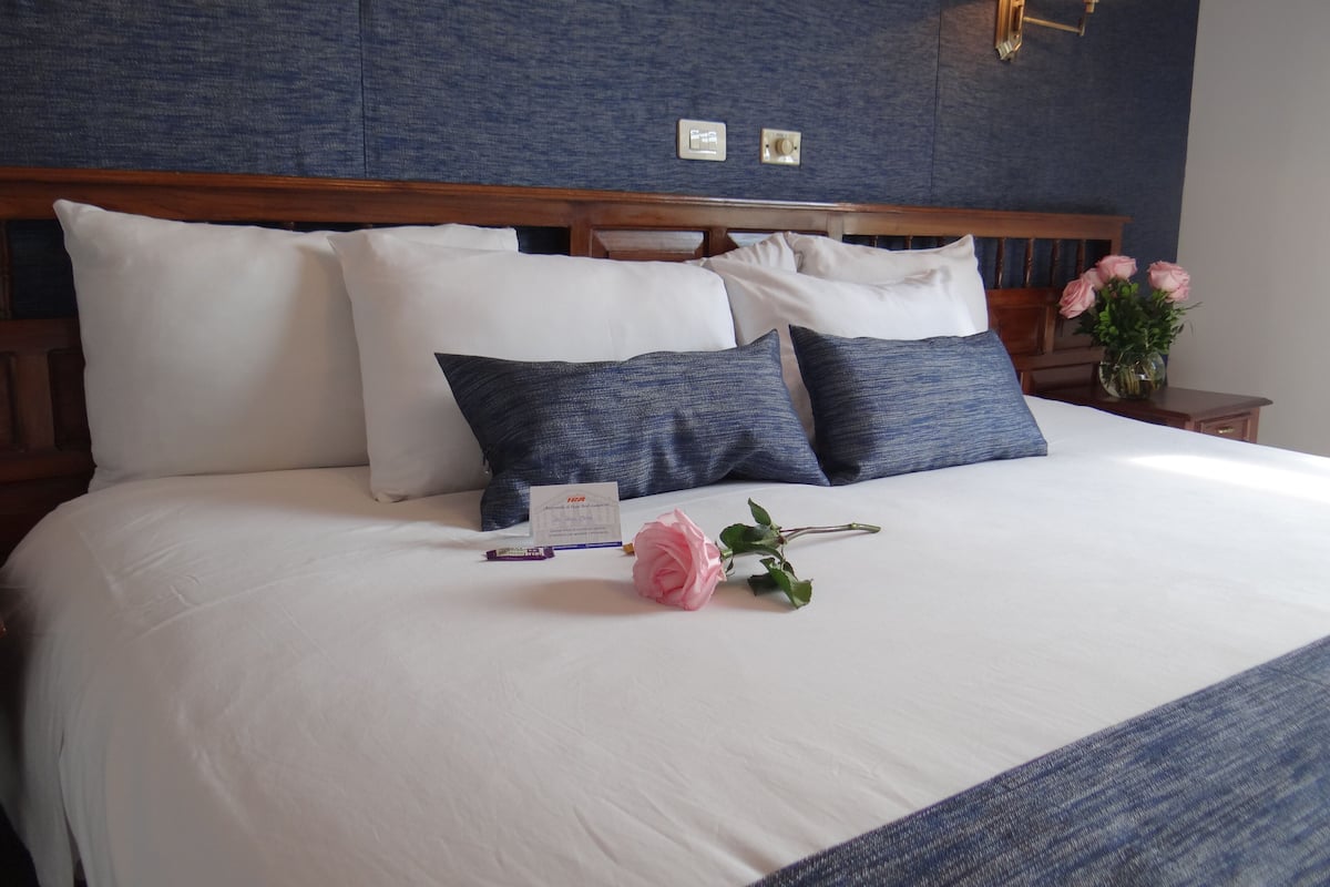 A neatly made bed with white and blue bedding, a bedside lamp, and a vase with pink flowers.