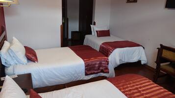 A hotel room with three single beds, each with white bedding and red accents. There is a wooden chair and a small table in the corner.