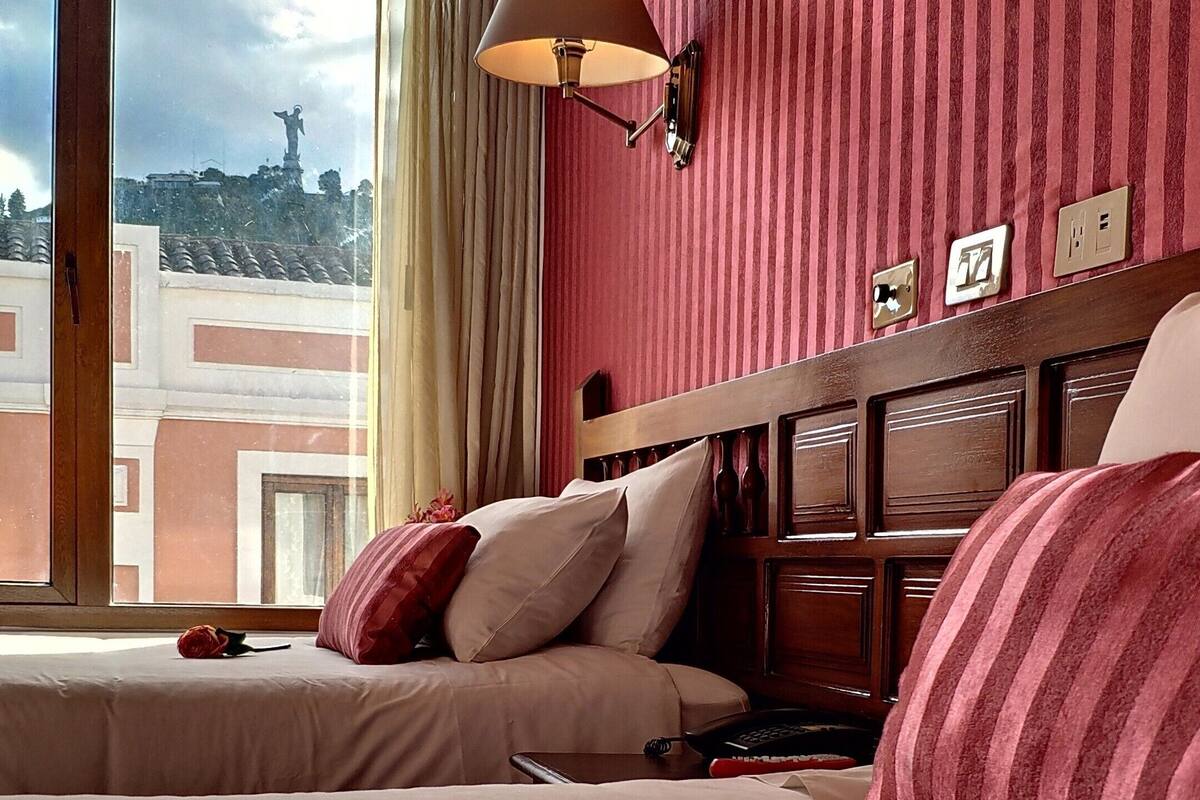 A hotel room with a large bed, a nightstand, a lamp, and a view of a cityscape through the window.