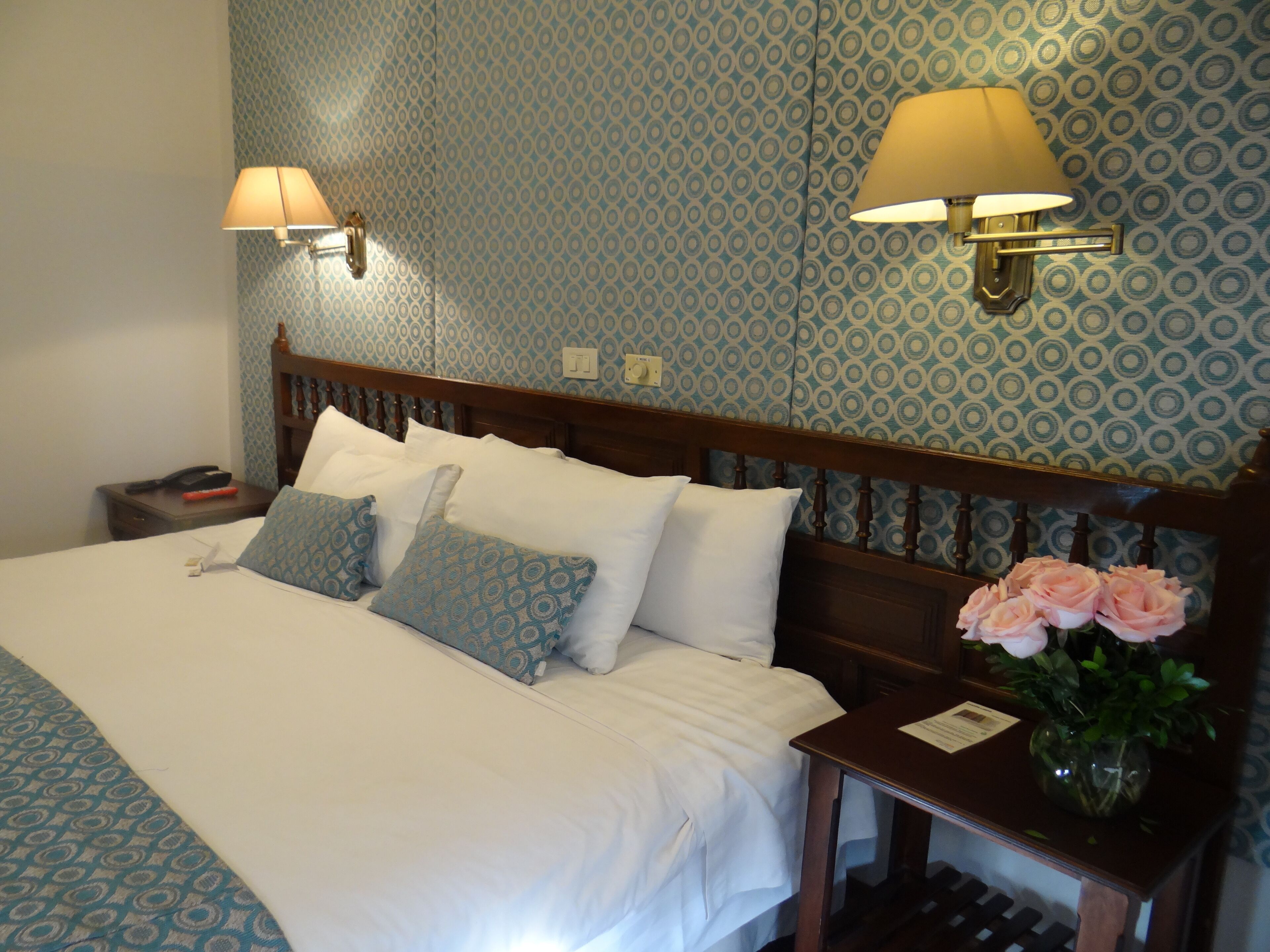 Una cama bien hecha con sábanas blancas y almohadas estampadas, un cabecero de madera, dos lámparas de noche, una mesita de noche con un teléfono y un libro, y un jarrón con flores sobre la mesita.