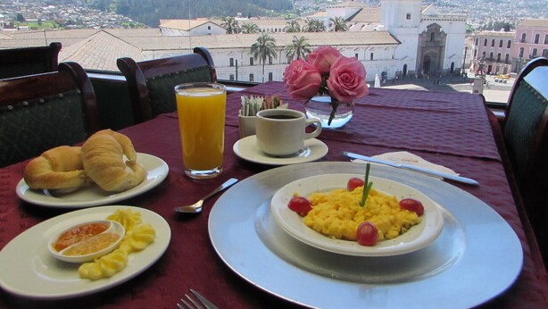 A table with breakfast items, a glass of orange juice, and a view of a cityscape with a prominent white building in the background. - Real Audiencia Hotel  (Quito)